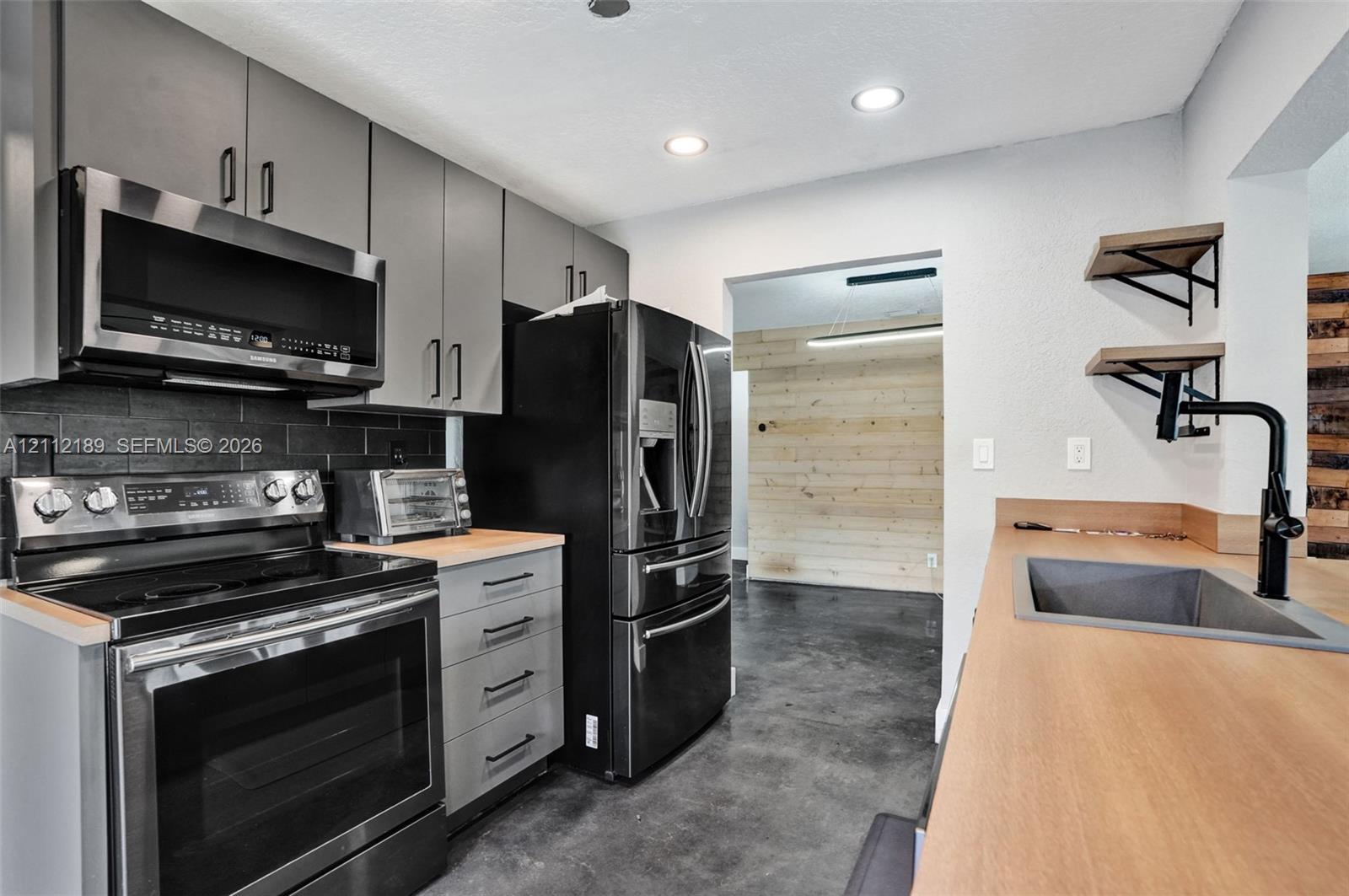 Kitchen featuring gray flat-panel cabinetry, dark brick-style backsplash, integrated stainless steel appliances, wood-finish countertops, and matte black plumbing fixtures