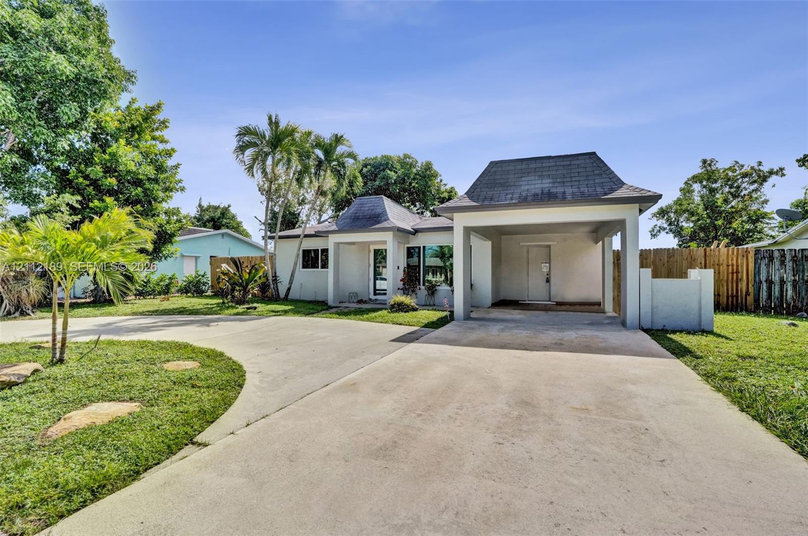 Single-story exterior featuring a hip roof, expansive concrete driveway, covered porte-cochre, and mature palm landscaping