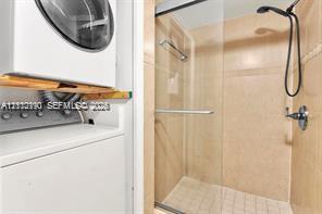 Shower stall featuring a frameless glass door, wall-mounted handheld showerhead, and tiled walls, accompanied by built-in laundry appliances with wood-finish shelving