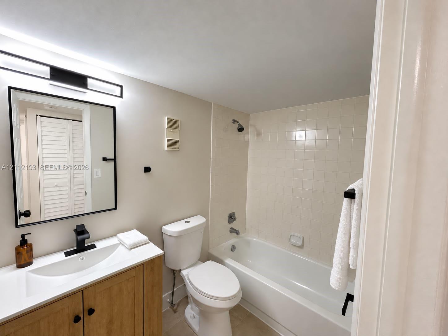 Bathroom featuring a wood-finish vanity with a white integrated sink, matte black faucet, rectangular framed mirror with integrated lighting, and a tiled shower-tub combination