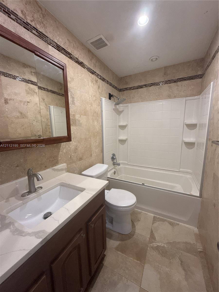 Bathroom featuring stone-finish wall and floor tiling, a vanity with a white countertop and rectangular undermount sink, a framed medicine cabinet, and a bathtub with a white surround