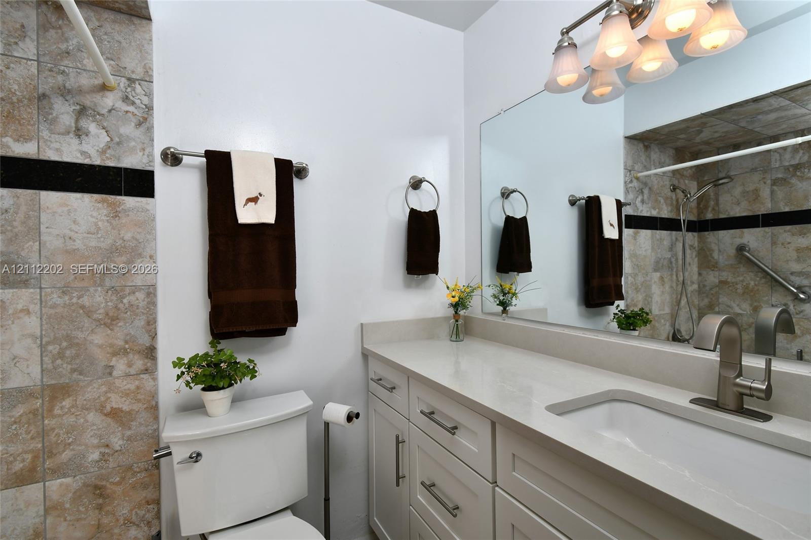 Bathroom vanity featuring a light-toned solid surface countertop, an integrated rectangular sink, brushed nickel hardware, and multi-drawer cabinetry, complemented by stone-tiled shower surround with decorative black accent tile