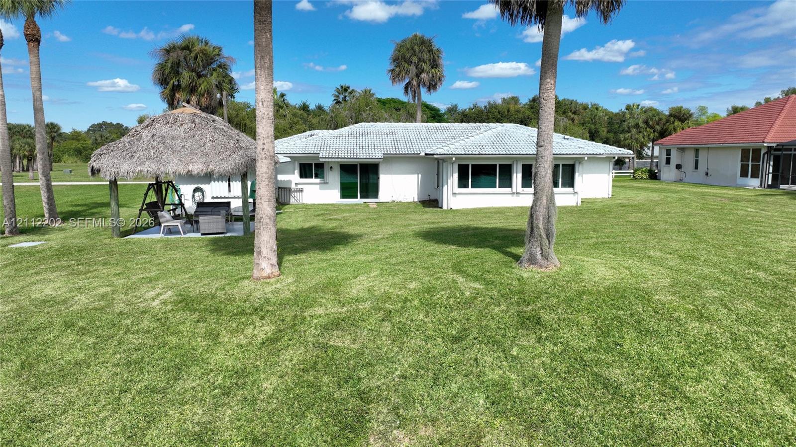 Rear exterior featuring a sprawling lawn, a detached thatched-roof palapa, and mature palm trees
