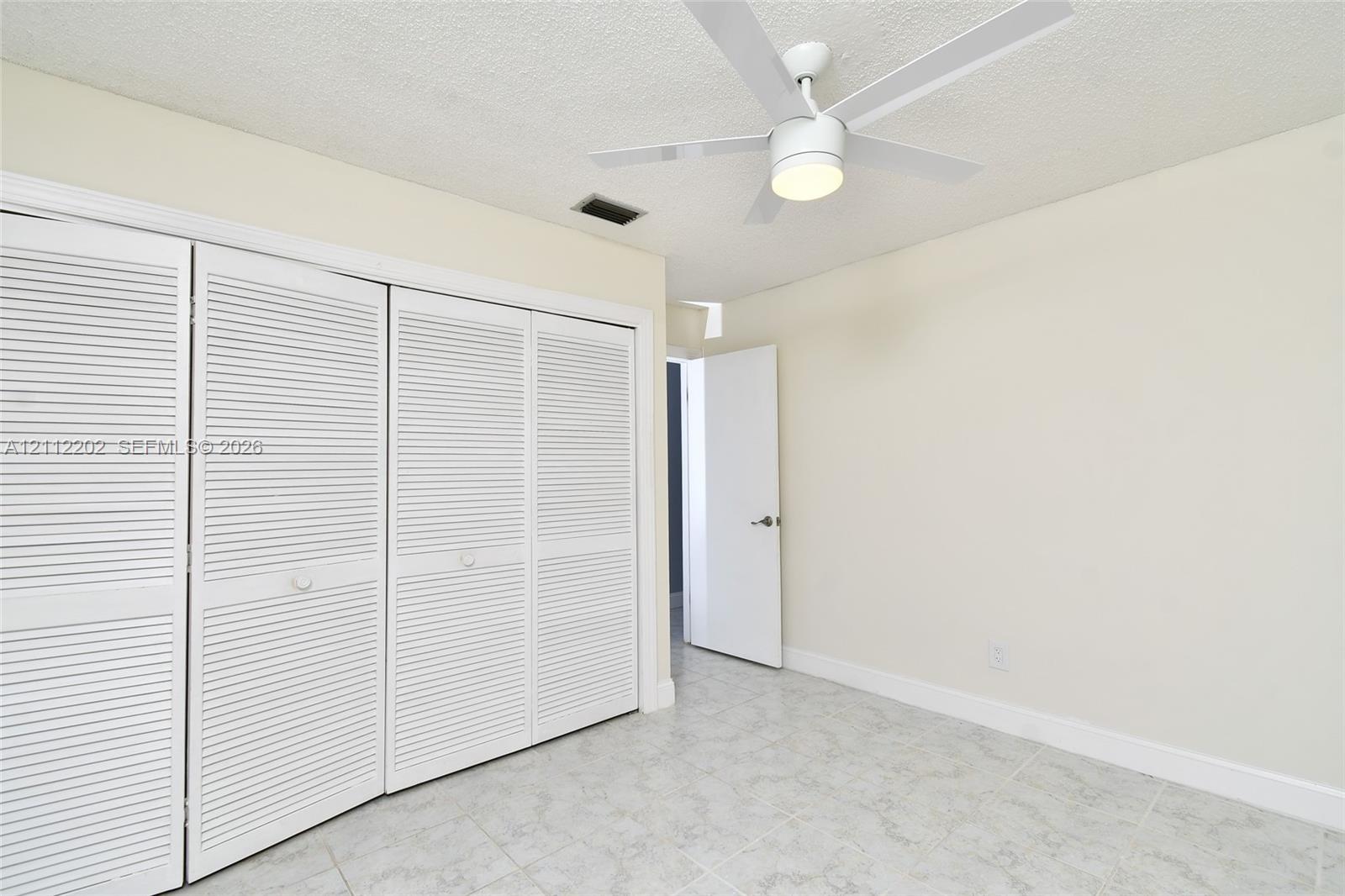Room featuring four-panel louvered closet doors, neutral wall finish, light-toned ceramic tile flooring, and a white ceiling fan with an integrated light fixture