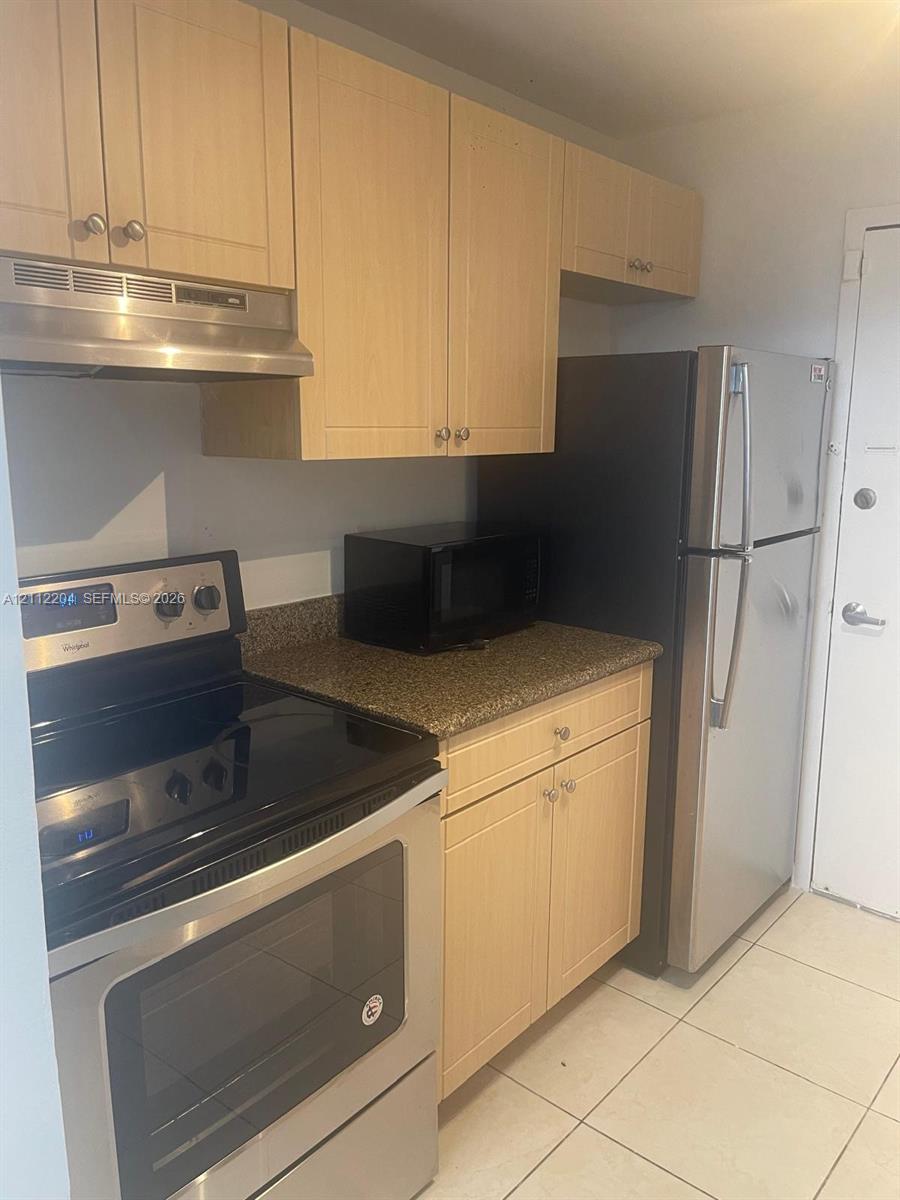Kitchen featuring light wood-finish cabinetry, stone-texture countertop, stainless steel range with overhead hood, and ceramic tile flooring