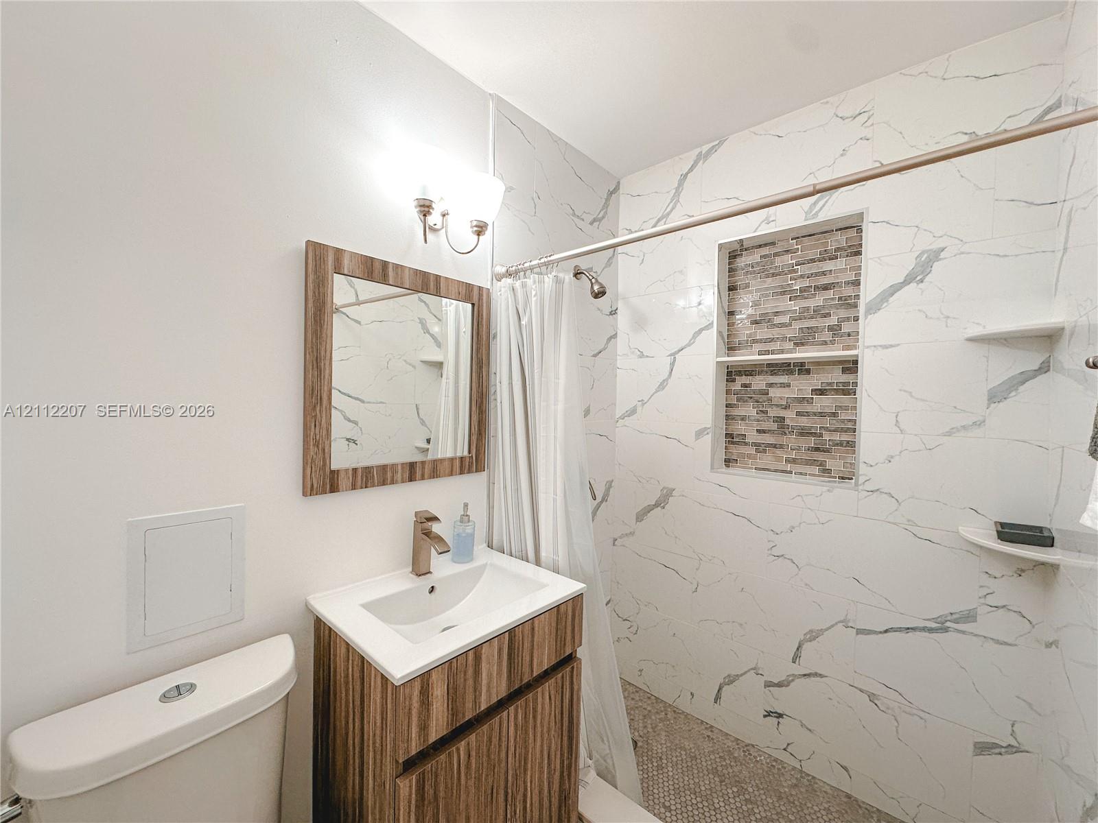 Bathroom featuring a vanity with a white countertop, wall-mounted light fixture, mirror with a wood-finish frame, and a shower area with large-format tile surround, stone-finish tile accent niche, and built-in shelving