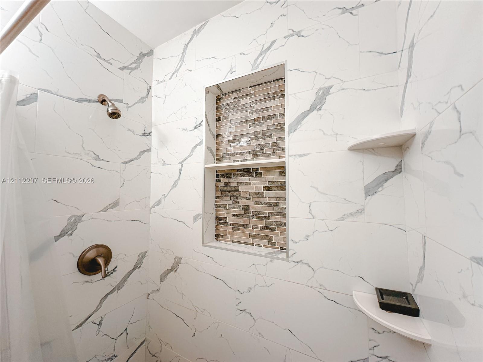Shower surround finished with large-format white veined tile, a multi-tier built-in tiled niche with decorative mosaic detailing, two integrated corner shelves, and a metal finish wall-mounted shower fixture