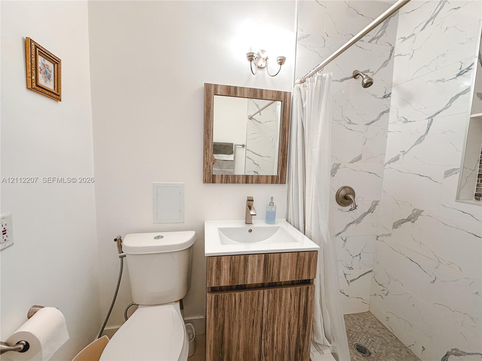 Bathroom featuring a wood-finish vanity with a rectangular sink, wall-mounted metallic lighting fixture, and shower enclosure with floor-to-ceiling stone-patterned wall tile