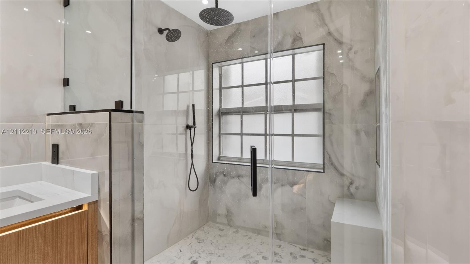 Walk-in shower featuring large-format stone-patterned tile, a rainfall showerhead with secondary handheld sprayer, a built-in bench, and a frameless glass enclosure