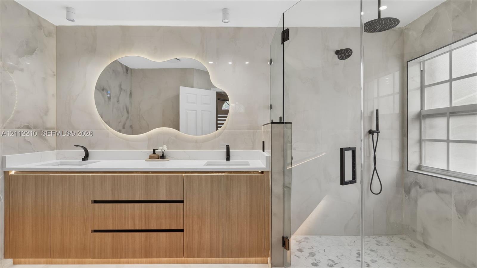 Floating wood-finish vanity with dual sinks, backlit organic-shaped mirror, glass-enclosed walk-in shower, and matte black fixtures