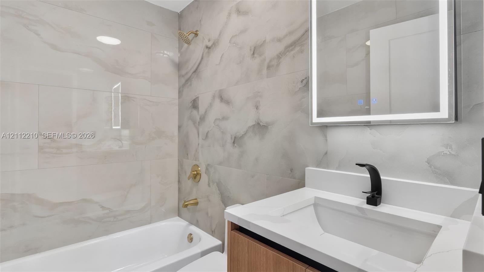Bathroom featuring large-format stone-pattern tile surround, wood-finish vanity with white countertop, integrated basin, matte black faucet, and LED-backlit mirror