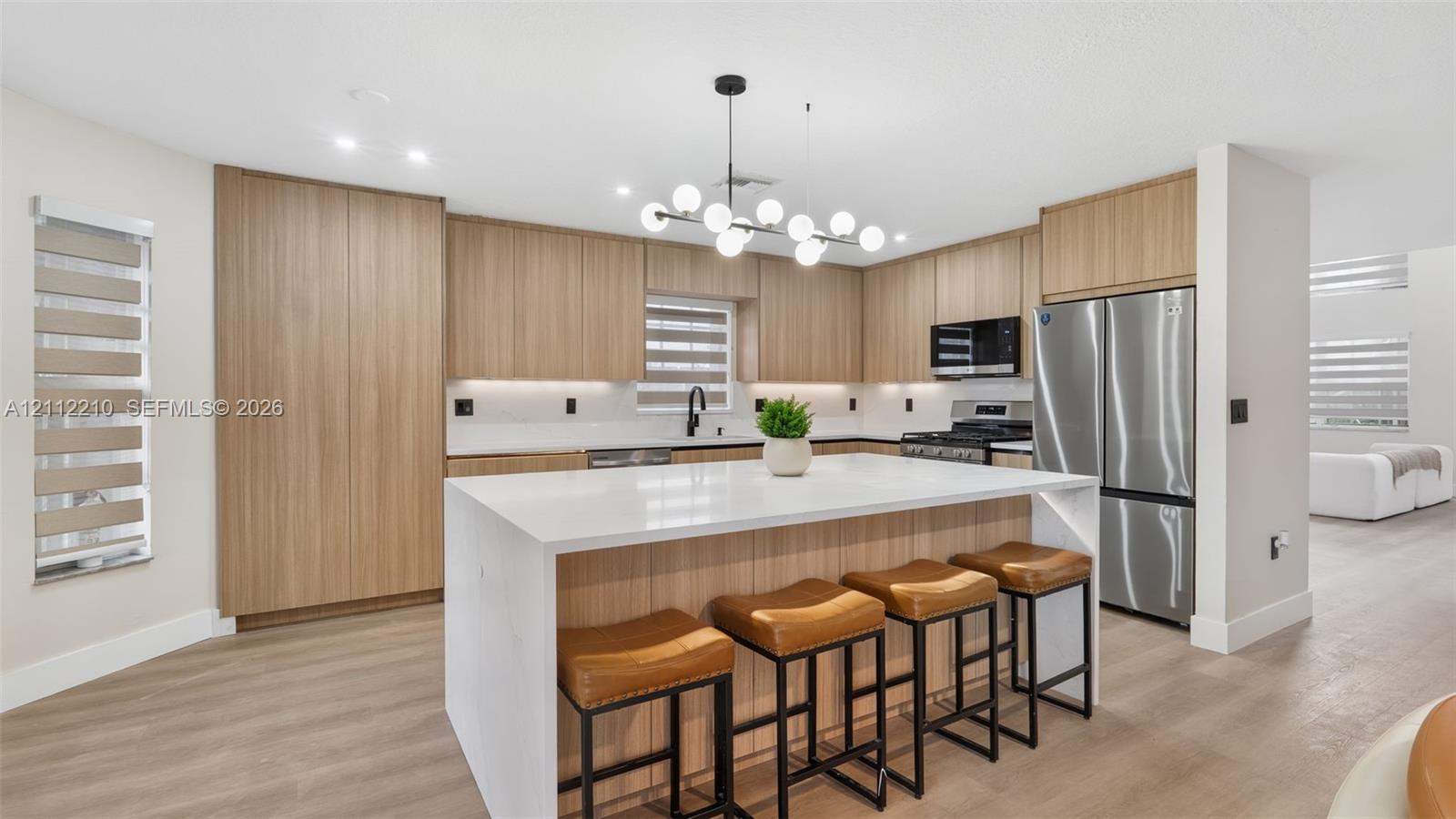 Kitchen featuring flat-panel wood-finish cabinetry, a large central island with waterfall-edge stone countertop, stainless steel appliances, and a modern multi-globe chandelier