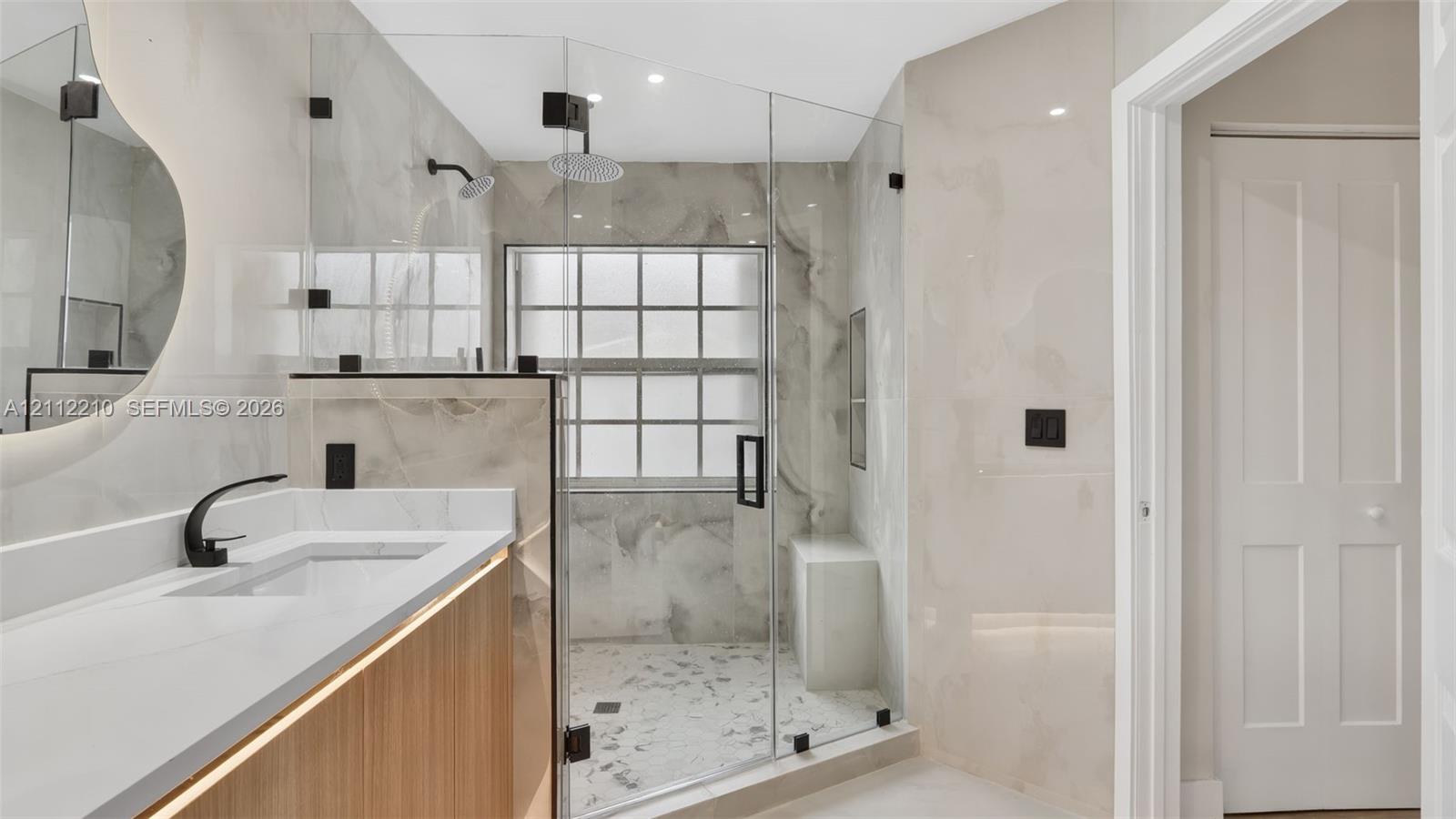 Bathroom featuring a wood-finish vanity with a quartz countertop, wall-to-wall stone-style tile, frameless glass shower enclosure with a built-in bench, rain-style showerhead, and matte black fixtures