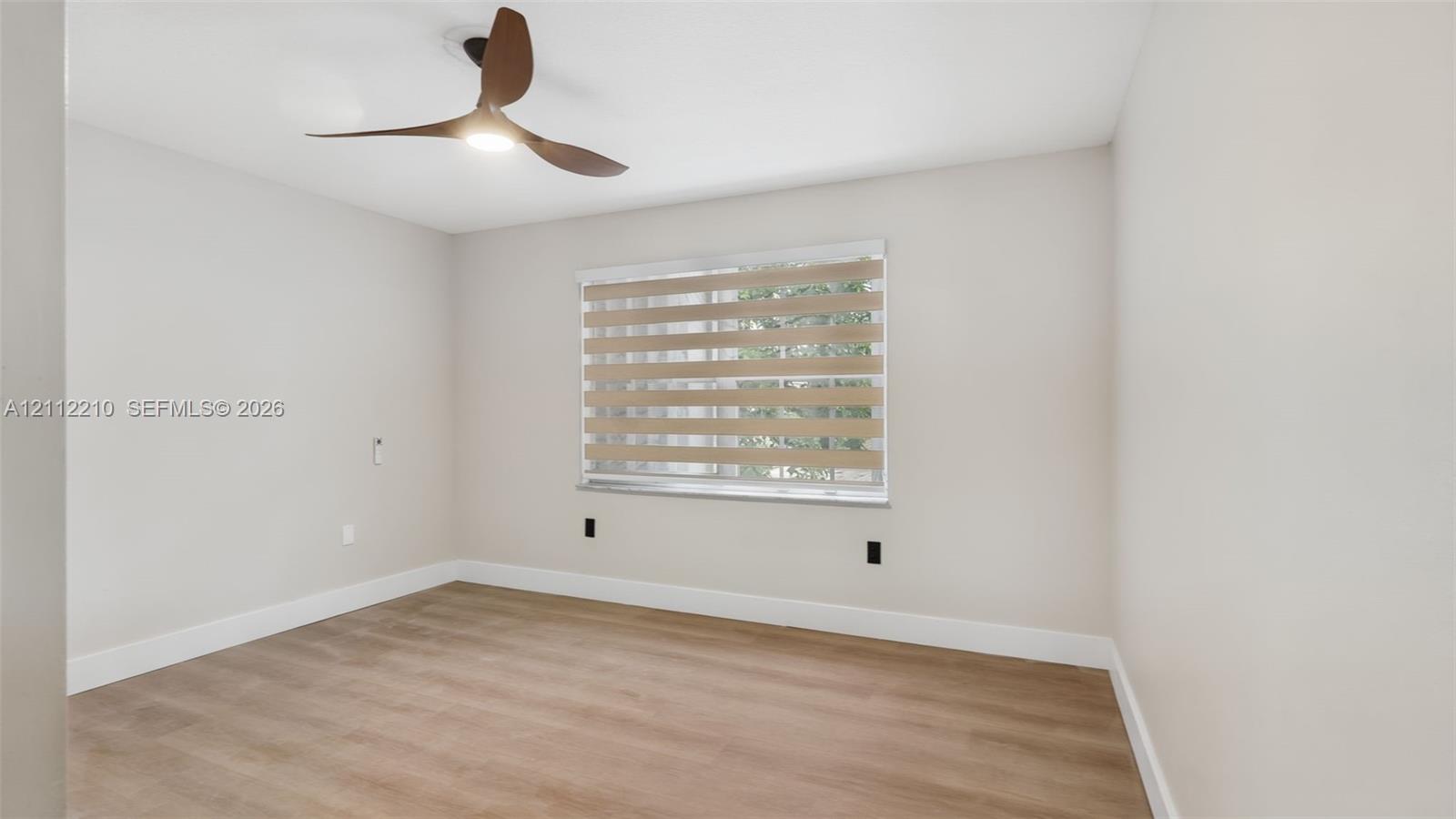 Neutral walls with wood-finish flooring, featuring a modern three-blade ceiling fan with integrated lighting and banded roller shades