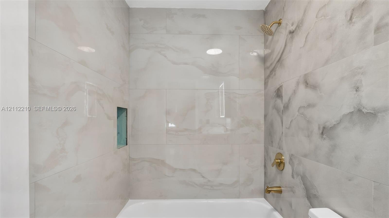 Bathtub surround featuring large-format stone-patterned tile, integrated recessed shelving, and brushed gold-tone shower fixtures