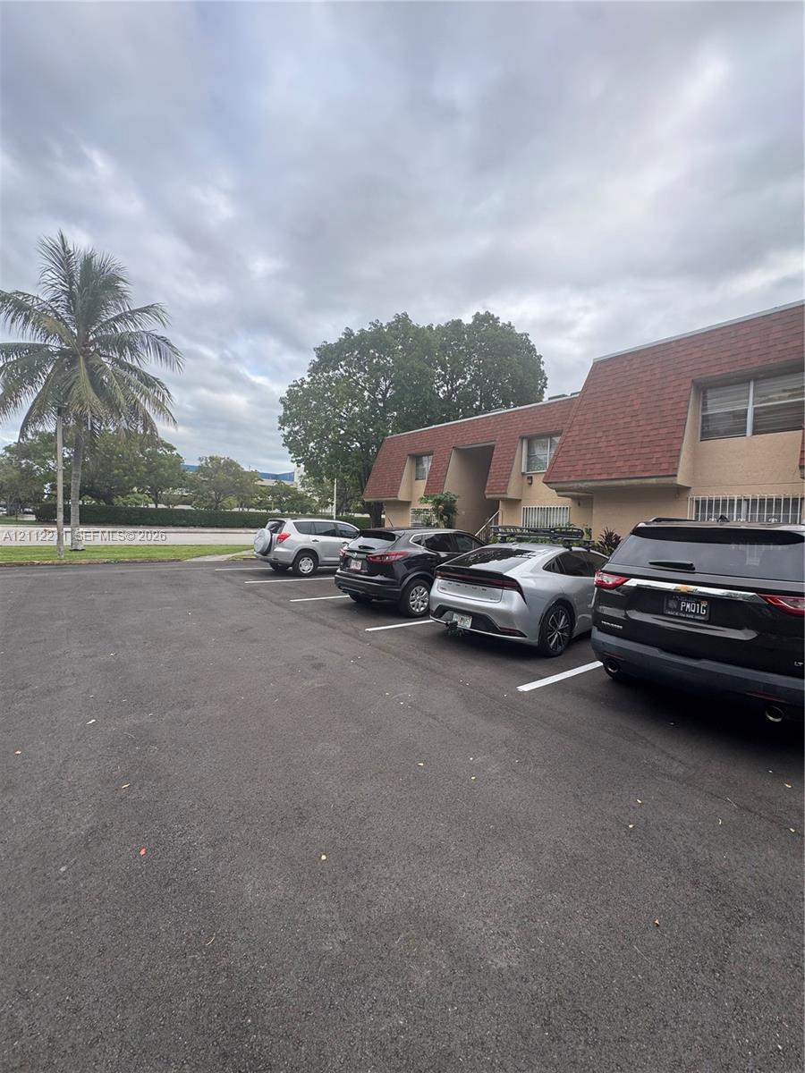 Asphalt parking area with painted stalls, featuring a multi-story exterior with textured siding, shingled mansard-style roofline, and established palm tree landscaping