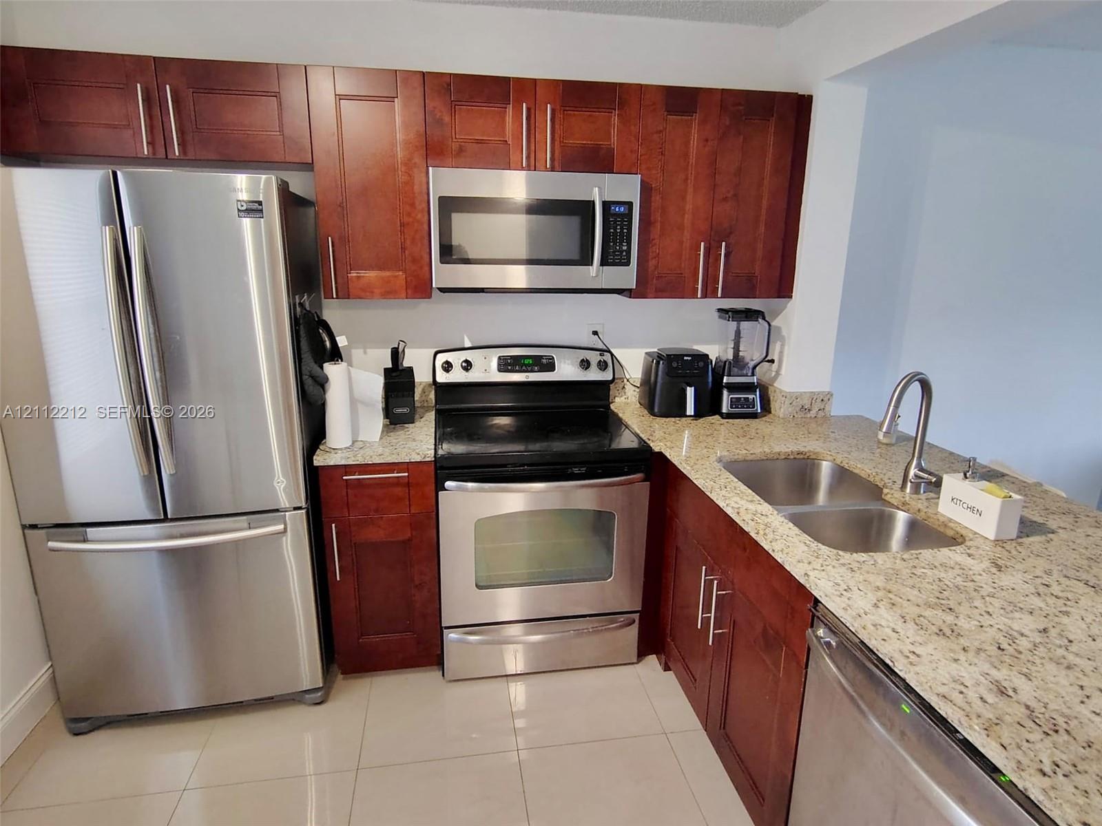 Kitchen featuring cherry-tone cabinetry, stone-finish countertops, stainless steel appliances, a dual-basin undermount sink, and light-tone tile flooring