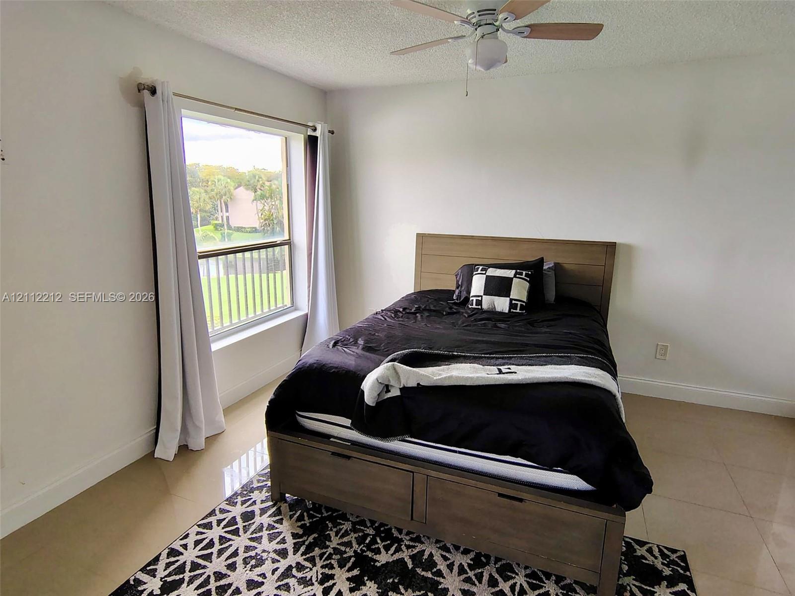 Neutral interior space featuring polished tile flooring, a textured ceiling, a ceiling fan with light fixture, and a large window with outdoor greenery exposure