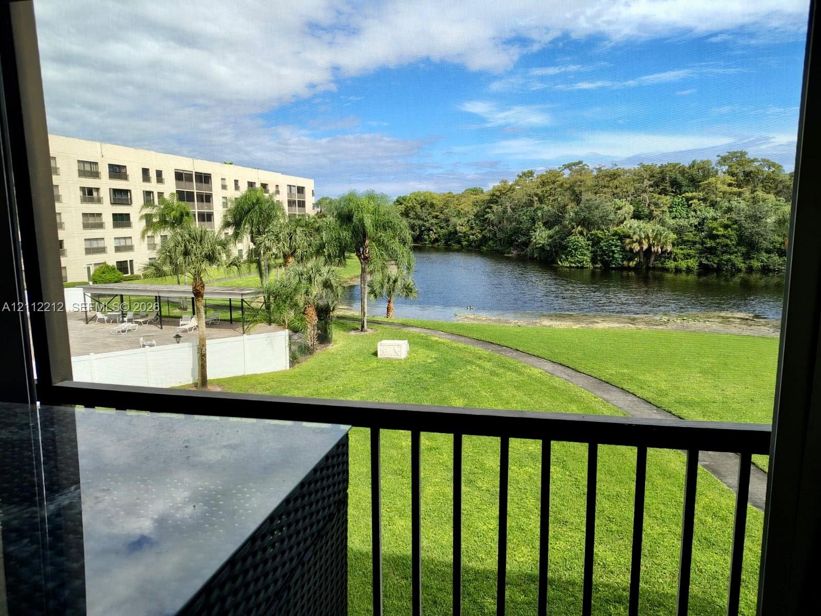 Private balcony with metal railing, stone-surface counter, lake frontage, manicured lawn, and mature palm trees