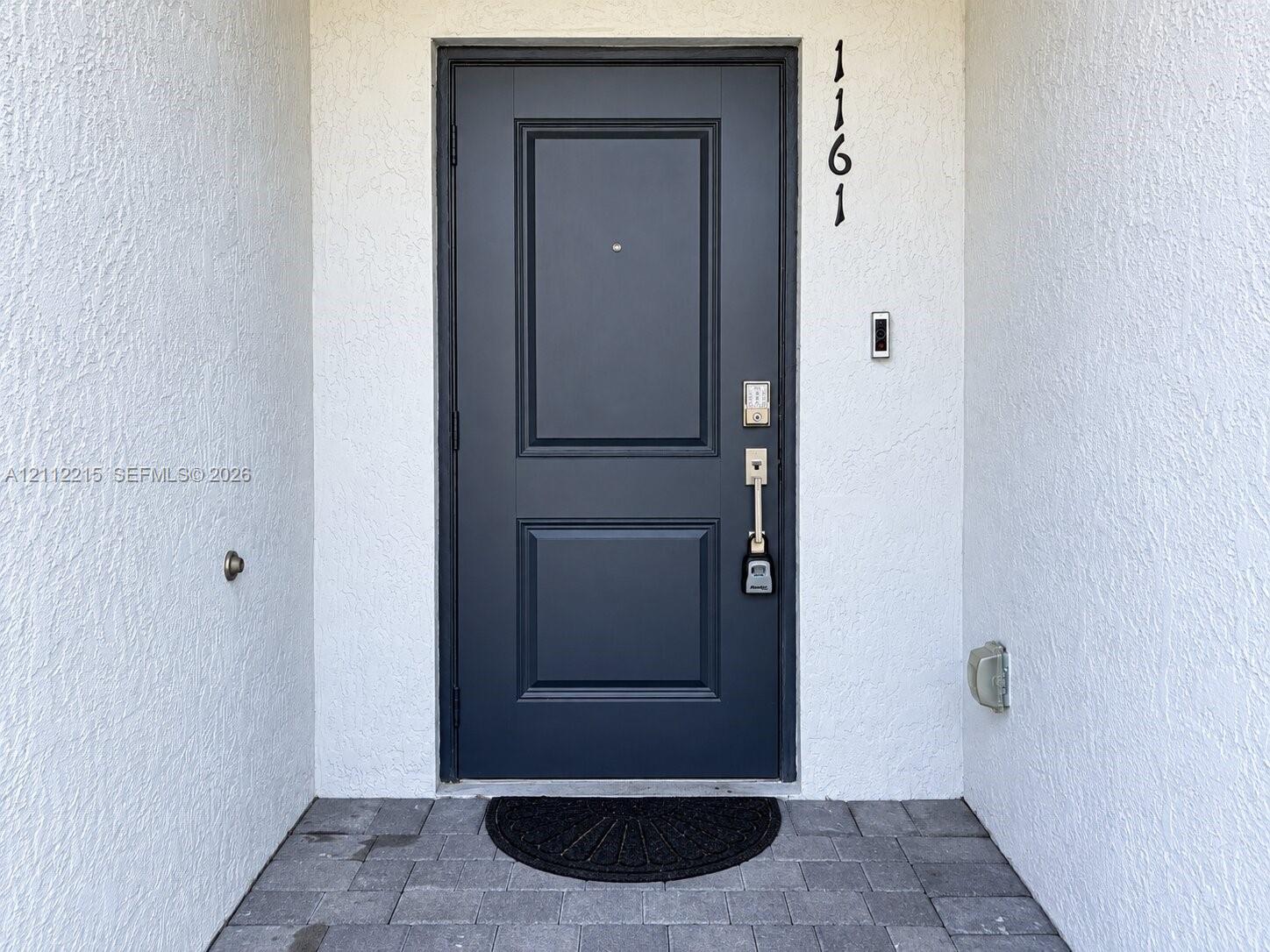 Dark-toned paneled entry door with a metallic peephole, matte-finish hardware, and exterior stone pavers