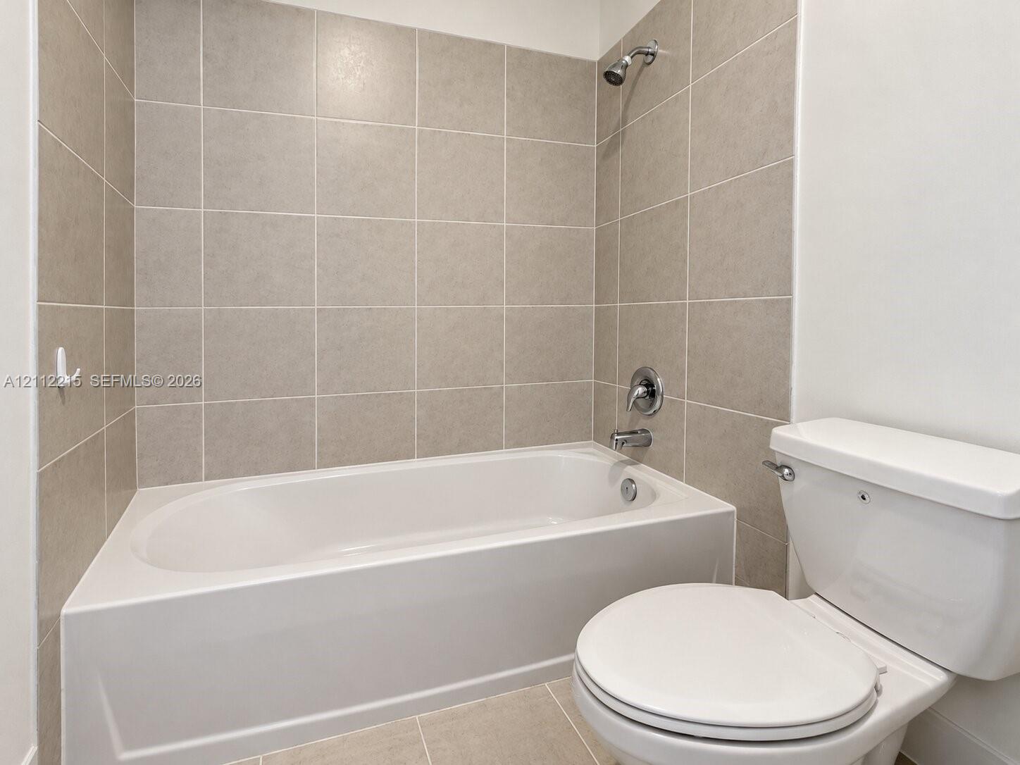 Bathroom featuring a composite soaking tub, a wall-mounted shower head with integrated diverter, neutral-toned square tile tub surround, and a porcelain commode