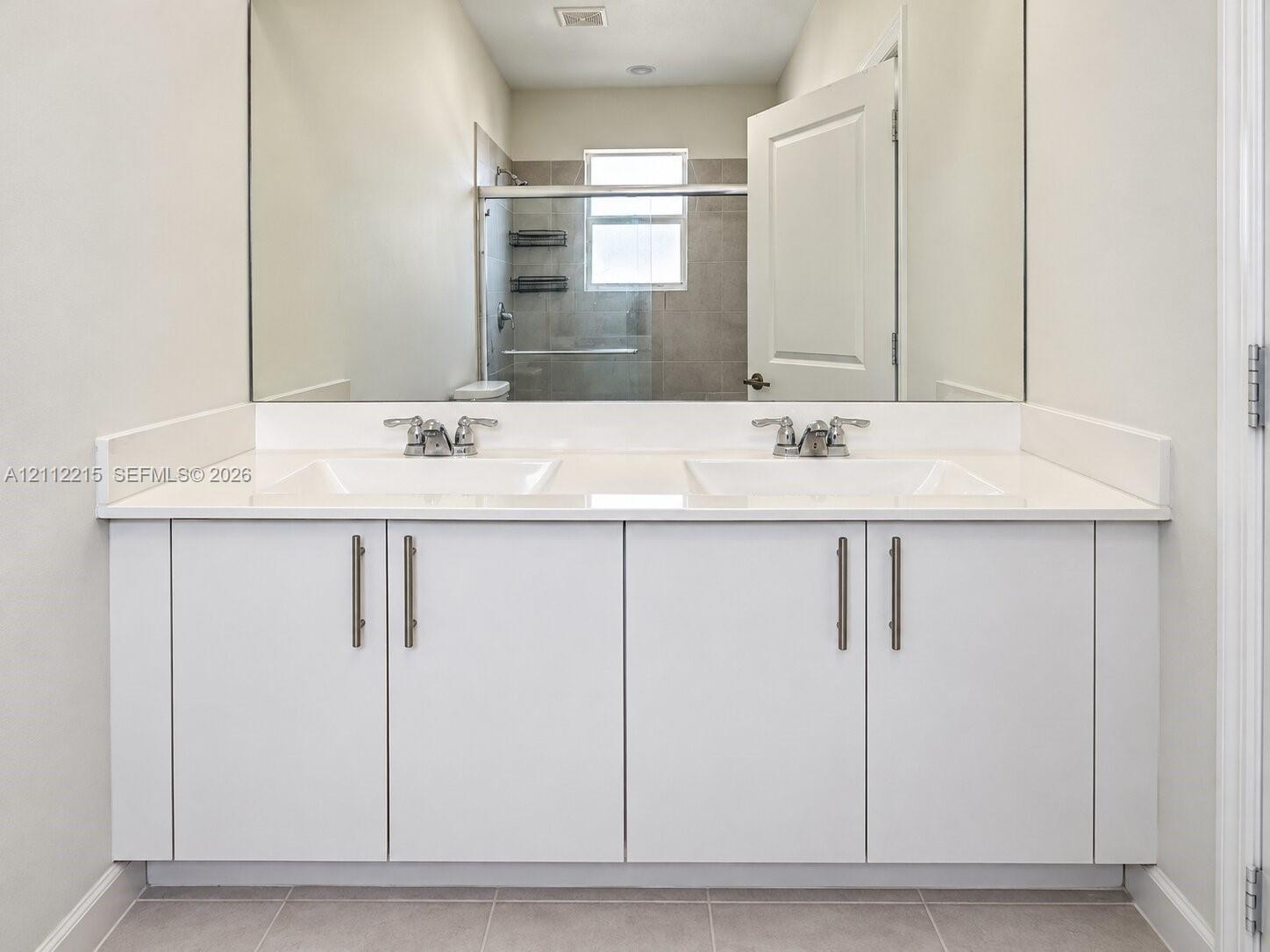 Dual-sink vanity featuring a solid surface countertop, white cabinetry with metallic hardware, and a full-width wall mirror