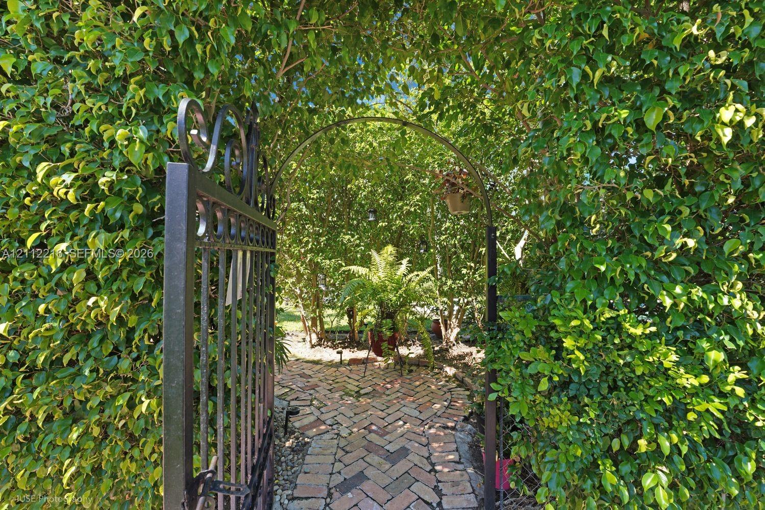 Metal gate with decorative scrollwork, arched entryway, and herringbone-pattern brick pathway lined with mature manicured hedging