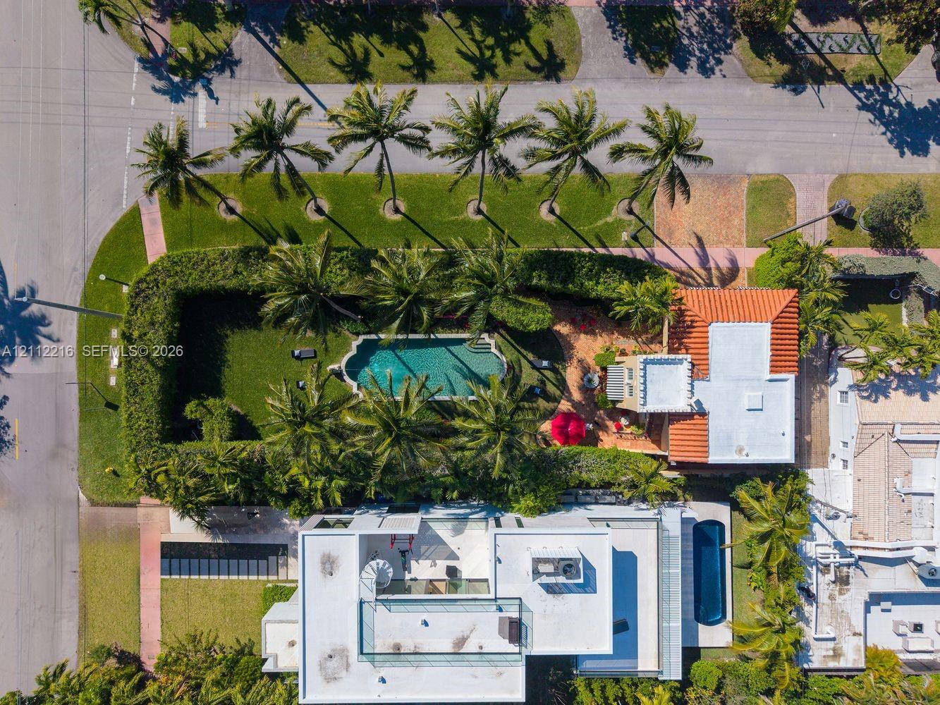 Aerial perspective of a multi-structure property featuring a swimming pool, a second elongated swimming pool, expansive manicured lawn, and mature perimeter landscaping