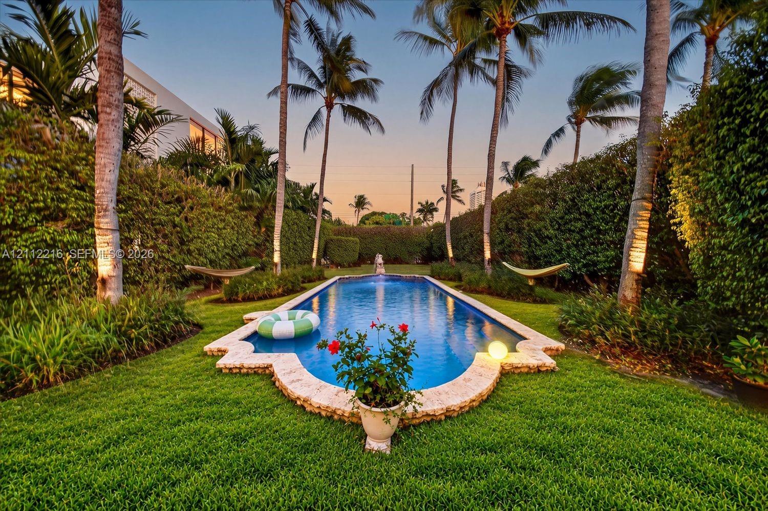 Rectangular swimming pool featuring a decorative stone coping, manicured lawn, perimeter hedge landscaping, and mature palm trees