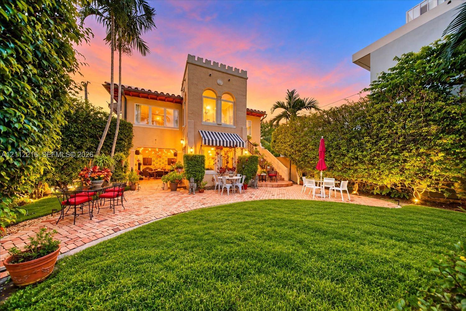 Mediterranean-inspired residence featuring a castellated tower, terracotta tile roof, brick-paved patio, manicured lawn, and perimeter privacy hedging