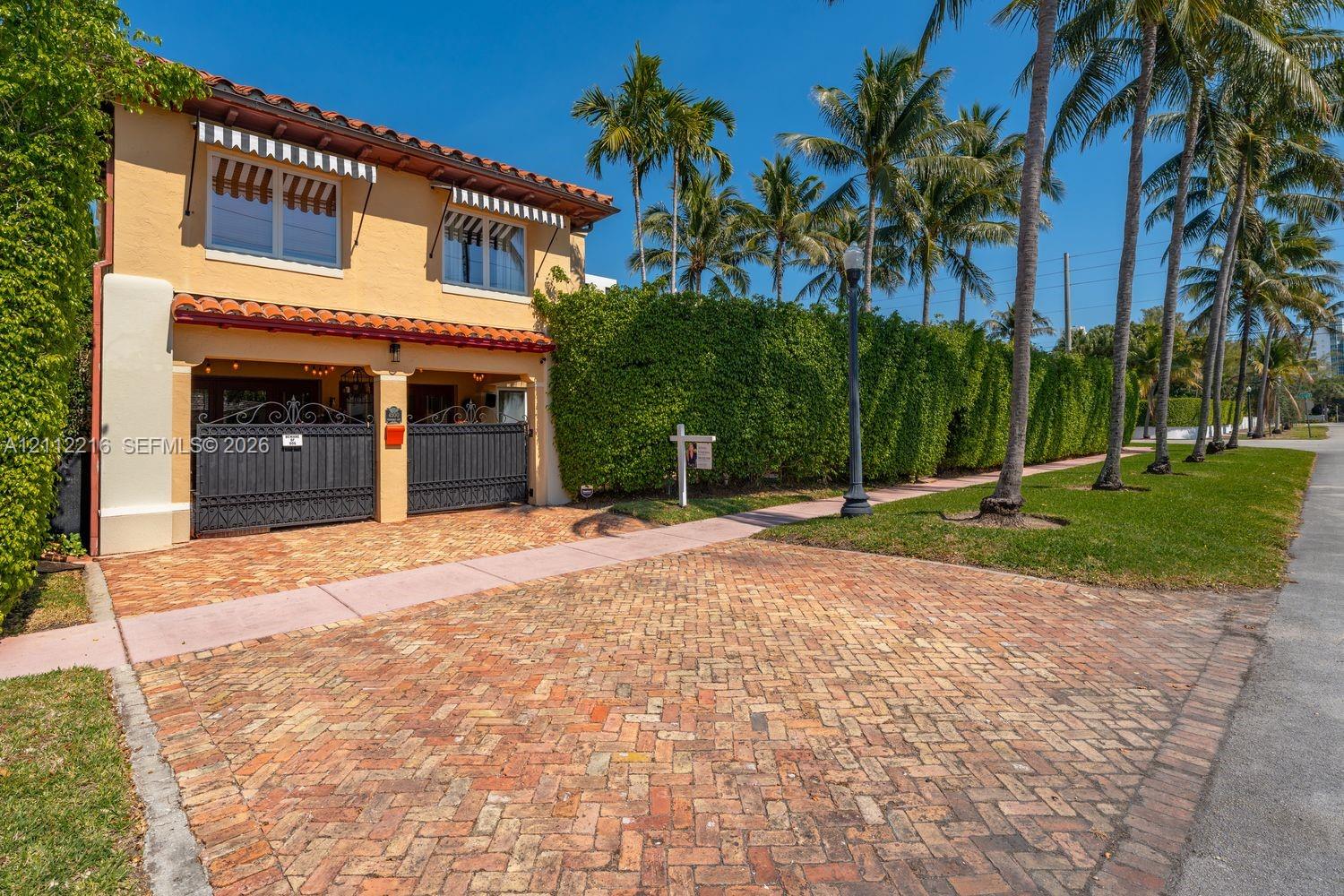Stucco exterior featuring a red tile roof, wrought-iron gate, striped window awnings, and herringbone-patterned brick driveway