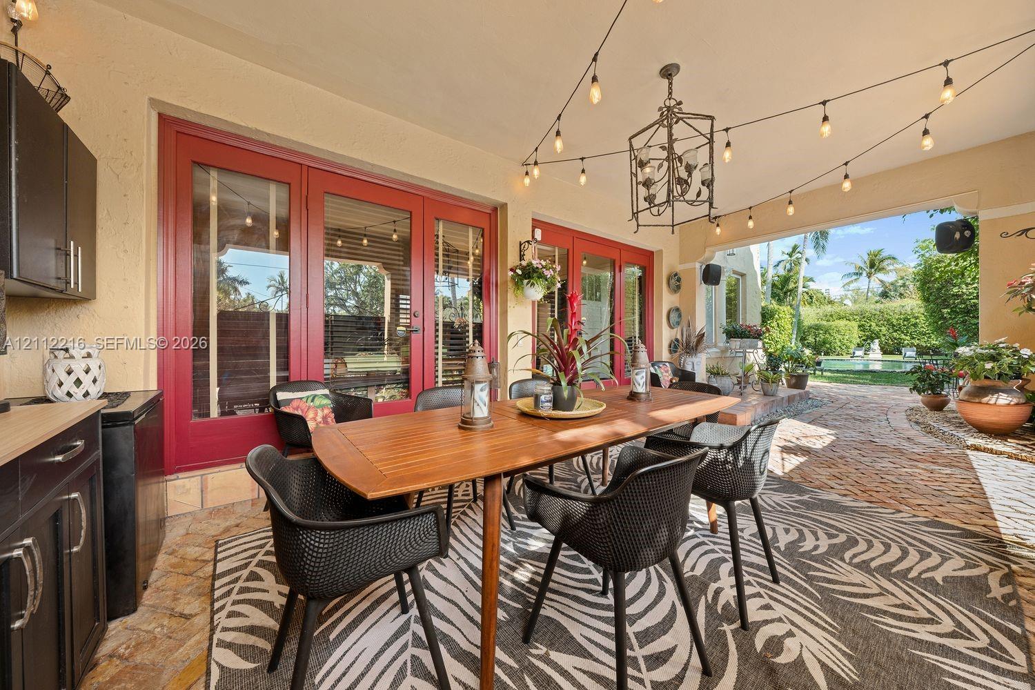 Covered patio featuring stone paver flooring, outdoor kitchen cabinetry, red-framed glass double doors, an overhead iron chandelier, and string lighting