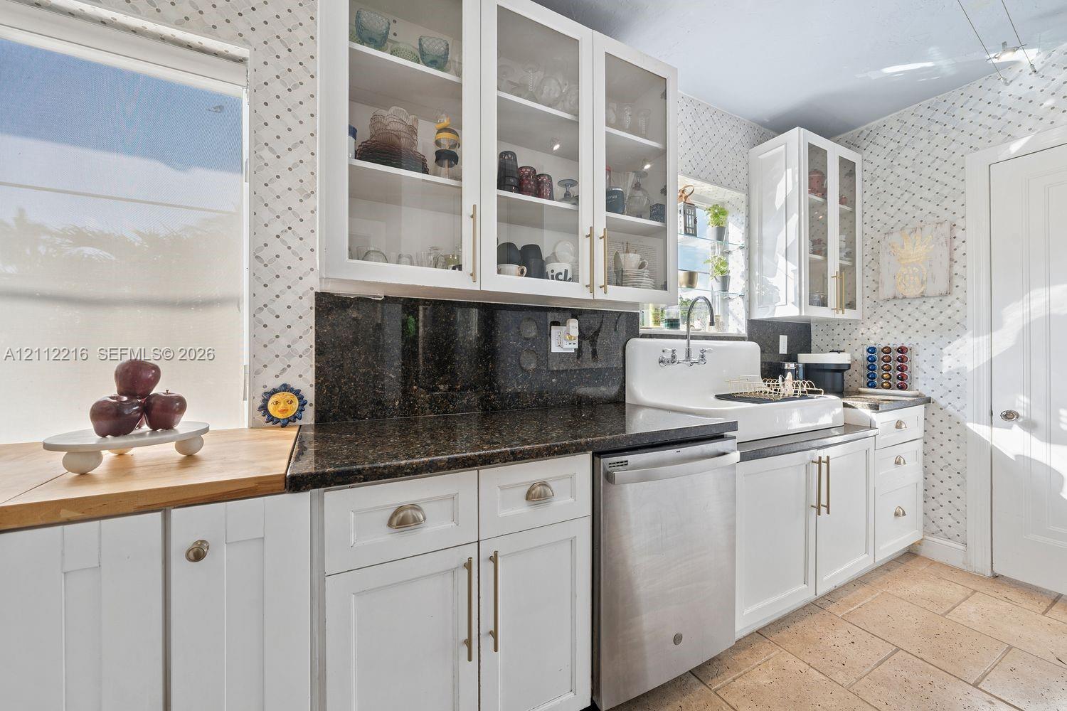 Kitchen featuring white cabinetry with glass-front uppers, dark stone countertops, stainless steel dishwasher, farmhouse-style sink with wall-mounted hardware, and stone tile flooring