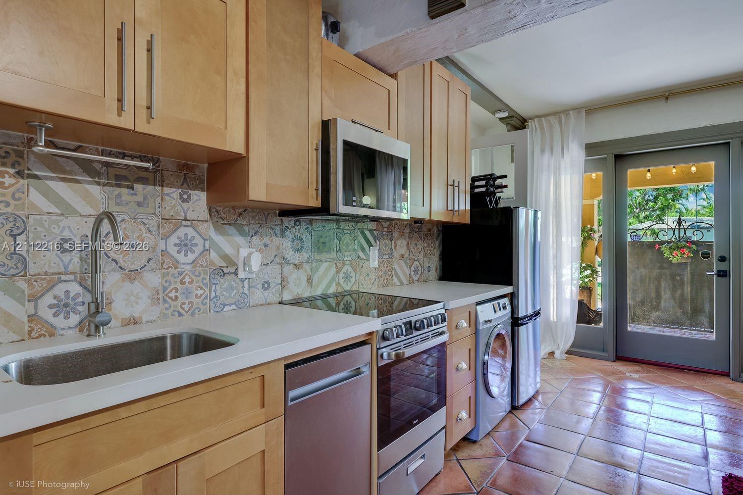 Kitchen featuring light wood cabinetry, patterned tile backsplash, solid surface countertops, stainless steel appliances, and terracotta tile flooring
