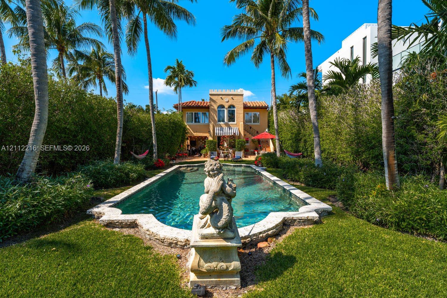 Custom in-ground swimming pool featuring a shaped stone-finished coping, central decorative sculpture, terracotta tile roof architecture, and mature perimeter landscaping