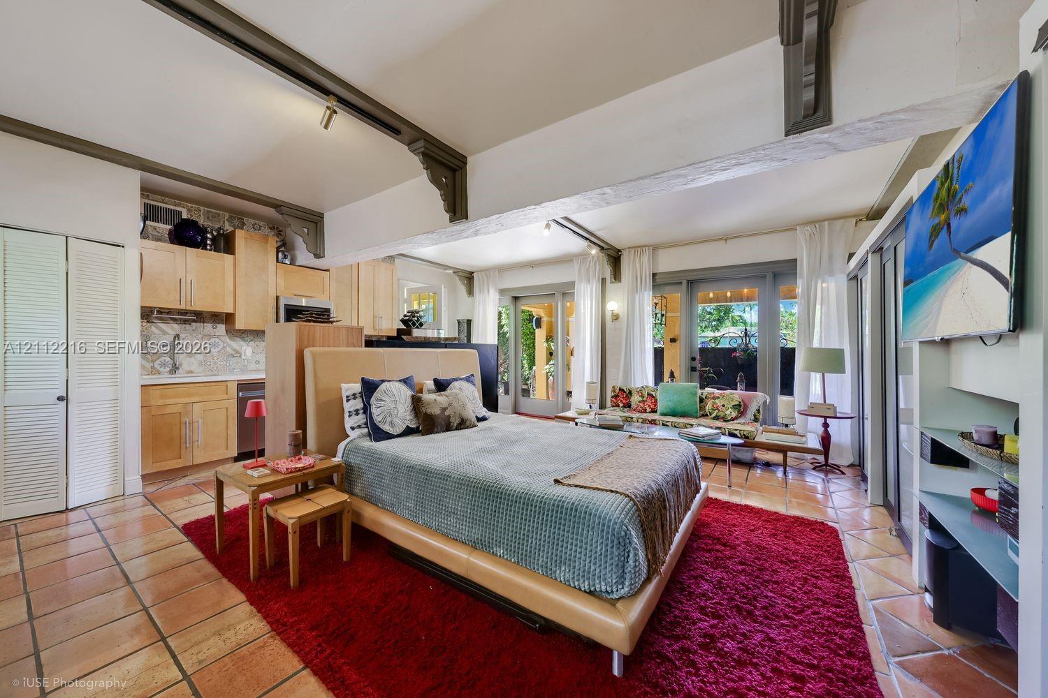 Open-concept living space featuring terracotta tile flooring, exposed structural beams with decorative corbels, natural wood cabinetry, patterned tile backsplash, and dual glass-paneled exterior doors