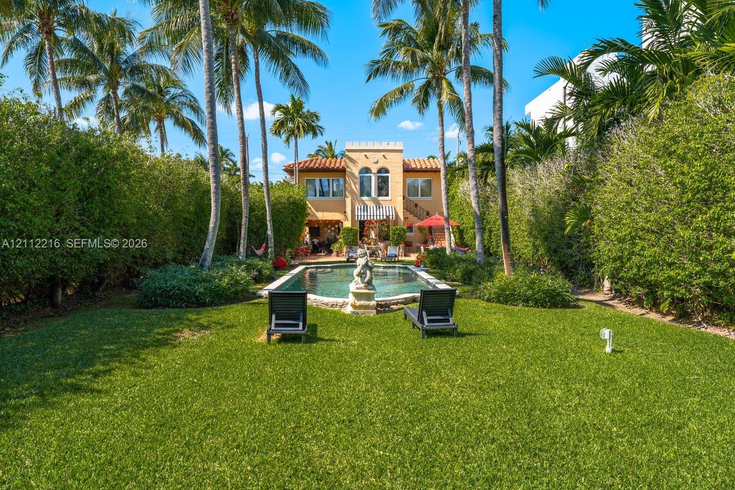 Expansive backyard featuring a rectangular swimming pool with a stone fountain, mature palm trees, and perimeter privacy hedging