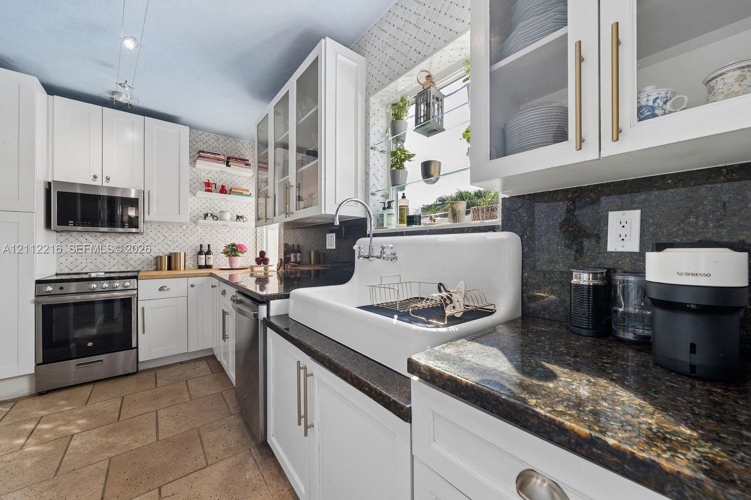 Kitchen featuring white cabinetry with glass-front uppers, apron-front farmhouse sink, stone countertops, stainless steel appliances, and patterned tile backsplash