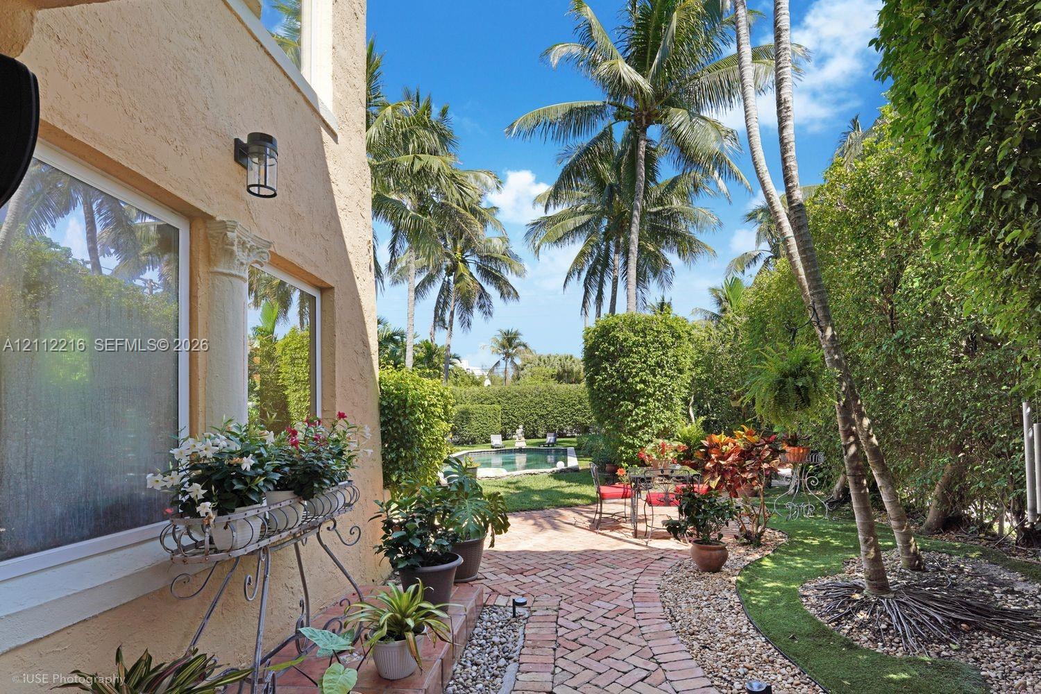 Stucco exterior with decorative column detail, brick-paved patio, lush landscaping with mature palm trees, and swimming pool