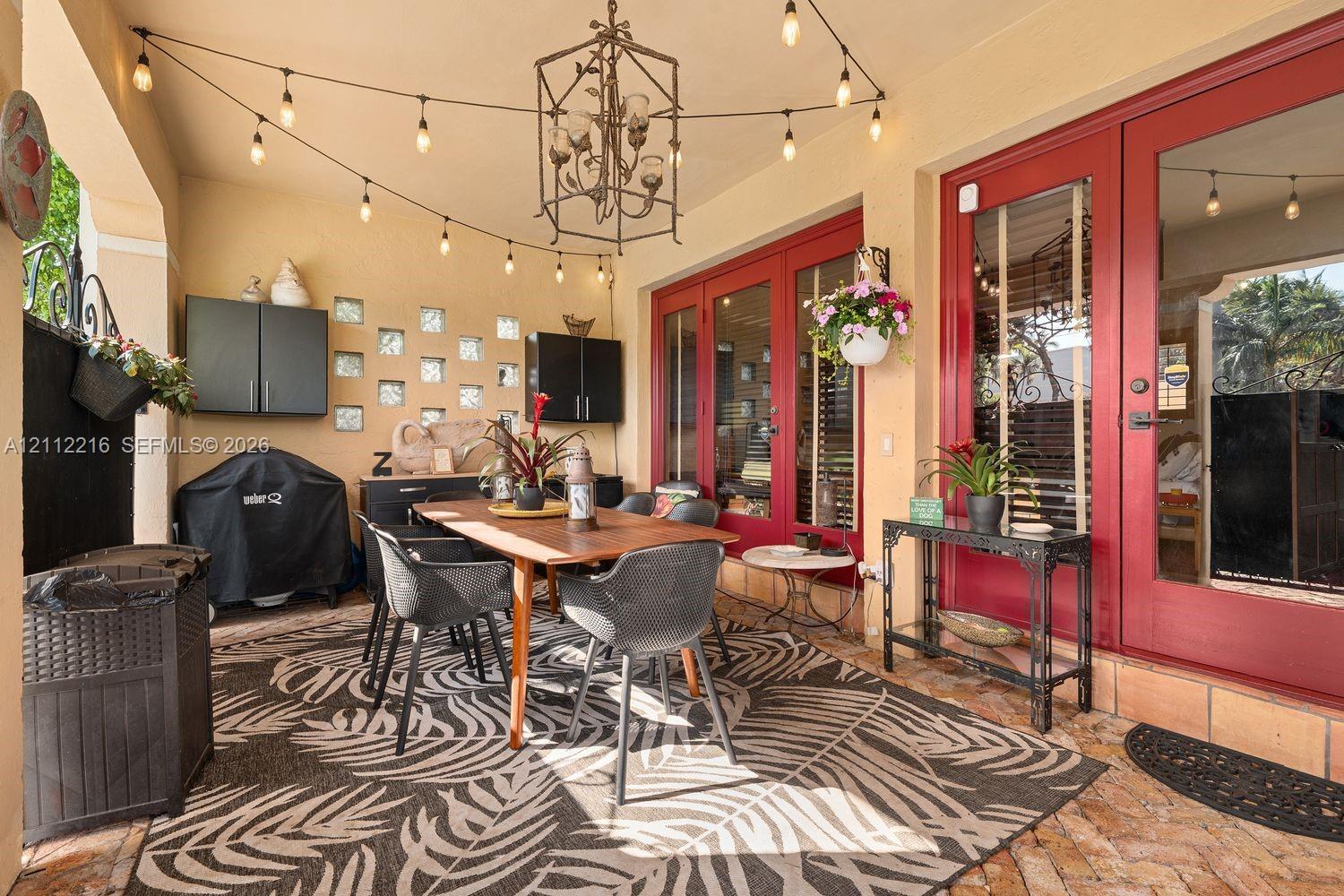 Covered patio featuring brick flooring, dual glass-paned doors with bold red frames, decorative glass block accents, and an overhead geometric chandelier