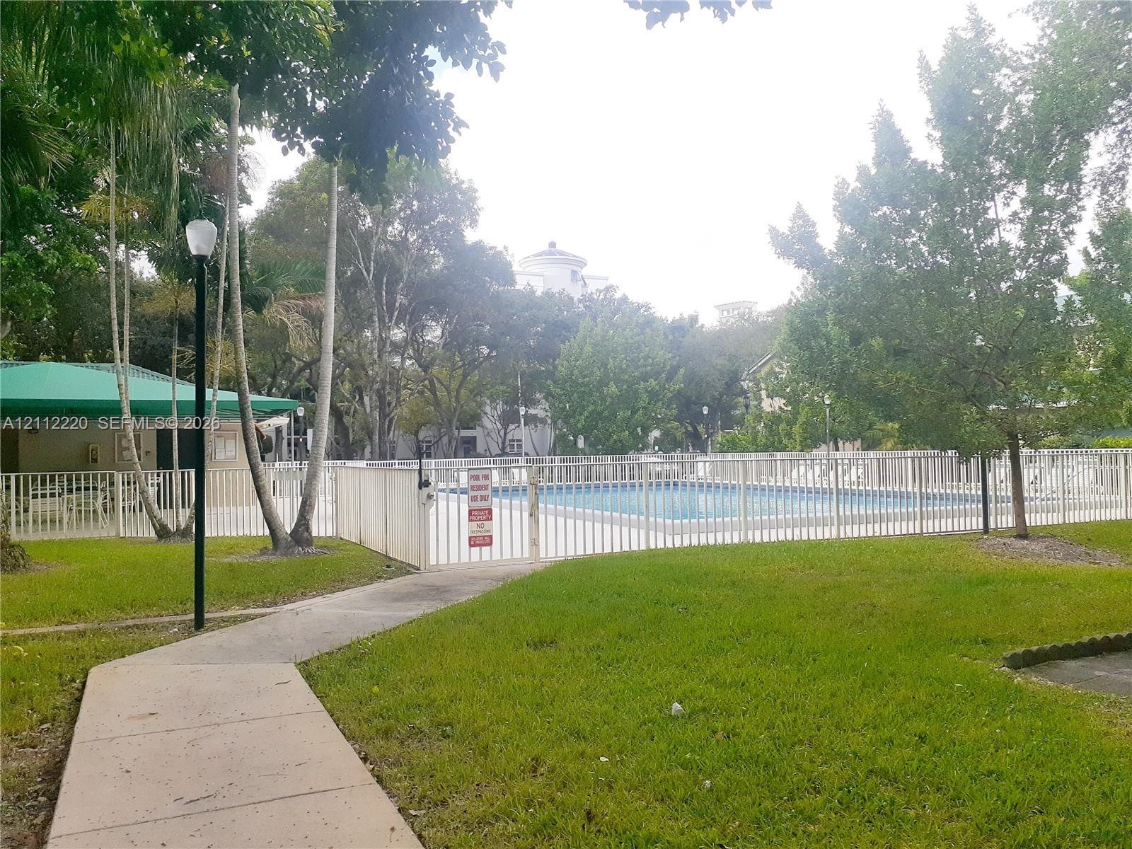 Swimming pool enclosed by white metal fencing, featuring an integrated cabana with a green canopy, concrete walkway, and mature landscaping