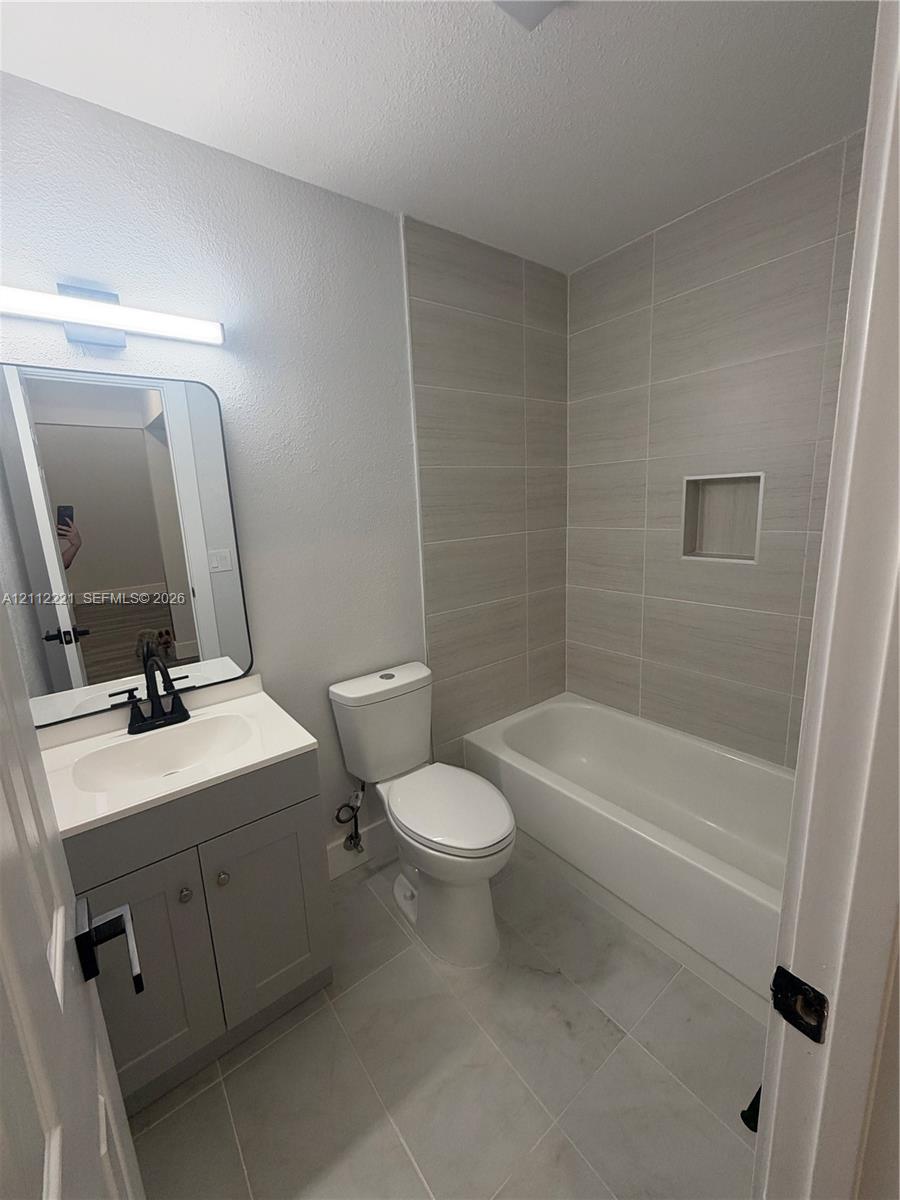 Bathroom featuring a gray shaker-style vanity with a white countertop, rectangular vessel sink with matte black faucet, wall-mounted mirror, and a tub-shower combination with floor-to-ceiling tile surround and built-in recessed shelving Bathroom featuring a gray shaker-style vanity with a white countertop, rectangular vessel sink with matte black faucet, wall-mounted mirror, and a tub-shower combination with floor-to-ceiling tile surround and built-in recessed shelving