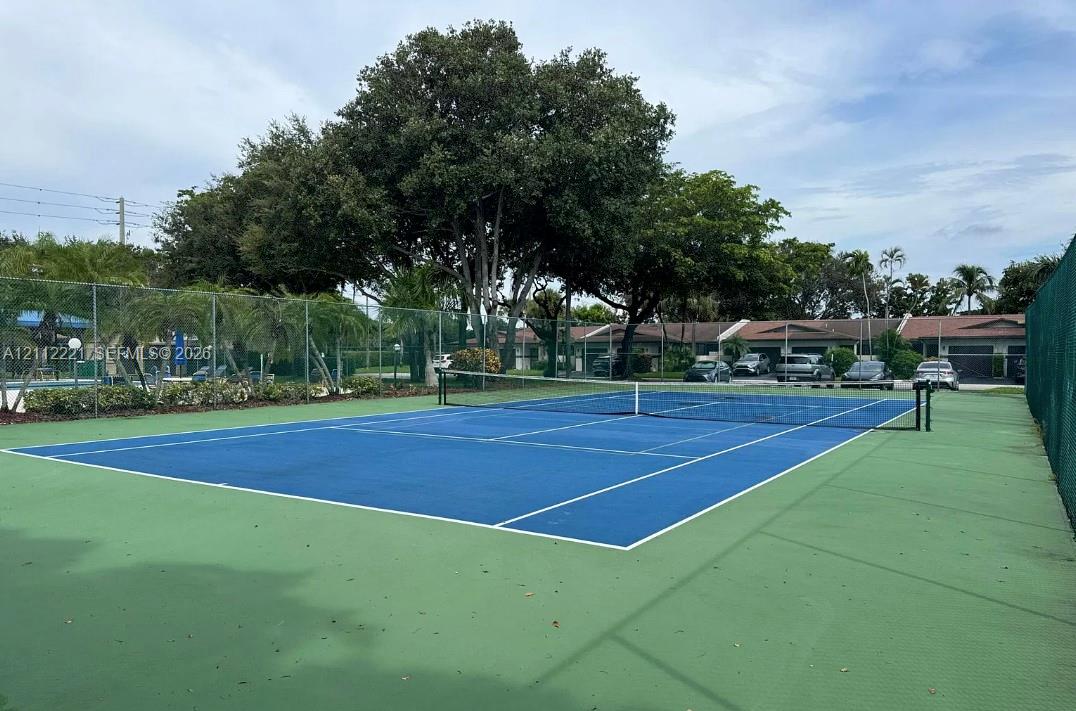 Tennis court with a dual-tone blue and green surface, enclosed by chain-link fencing and bordered by mature trees