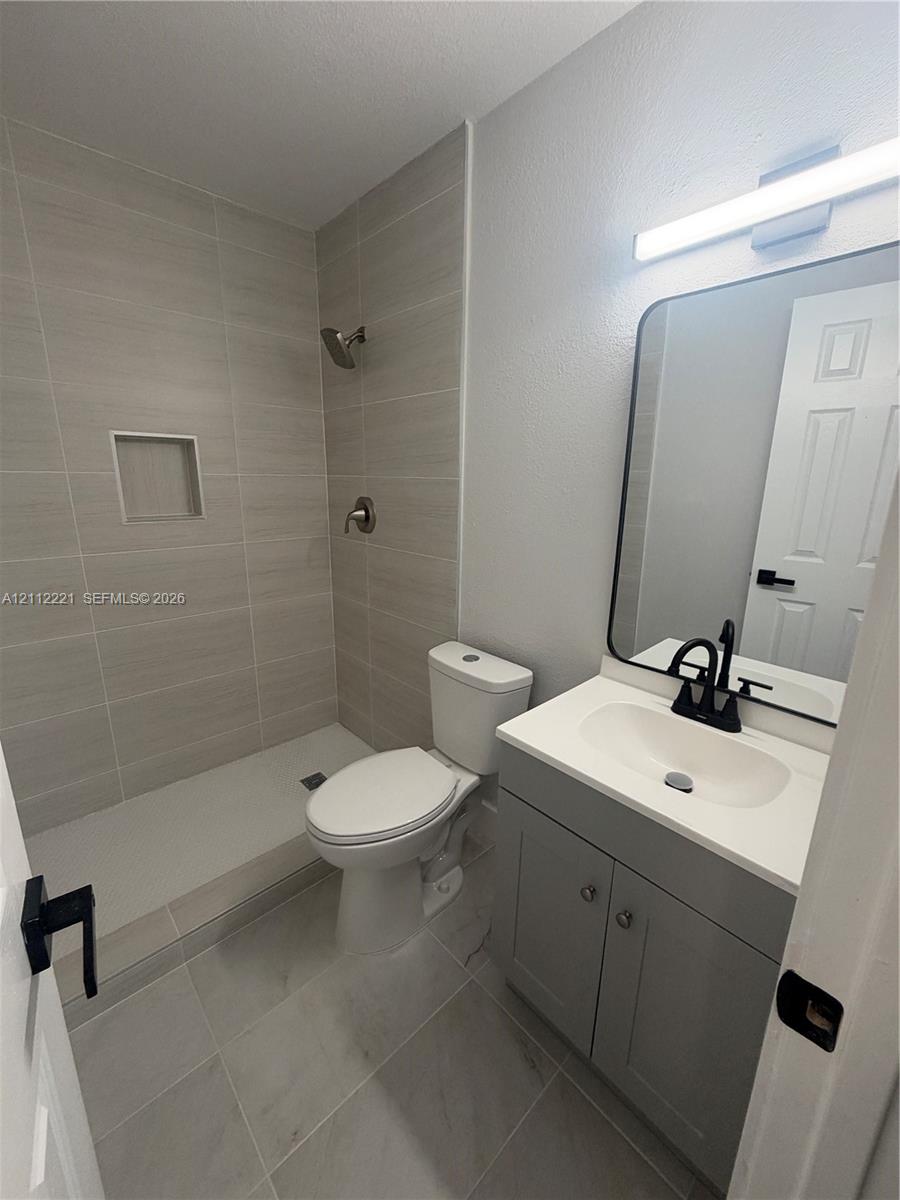 Bathroom featuring a walk-in shower with light-toned horizontal tile, a built-in recessed niche, a grey vanity with a white countertop, a rectangular mirror with a black frame, and matte black fixtures Bathroom featuring a walk-in shower with light-toned horizontal tile, a built-in recessed niche, a grey vanity with a white countertop, a rectangular mirror with a black frame, and matte black fixtures