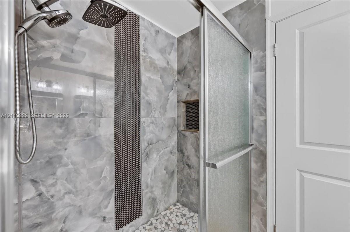 Walk-in shower featuring large-format wall tile with a contrasting mosaic accent strip, rain-style shower head, handheld sprayer, pebble-style shower floor, and a recessed wall niche
