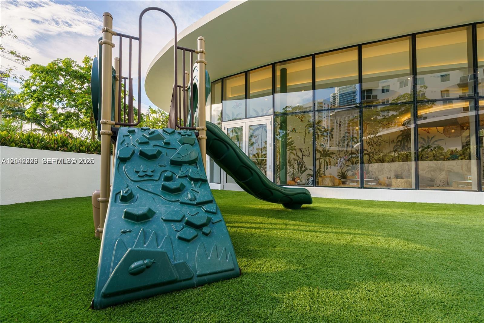 Outdoor recreation area featuring artificial turf, a permanent slide structure, floor-to-ceiling glass paneling, and white perimeter walling