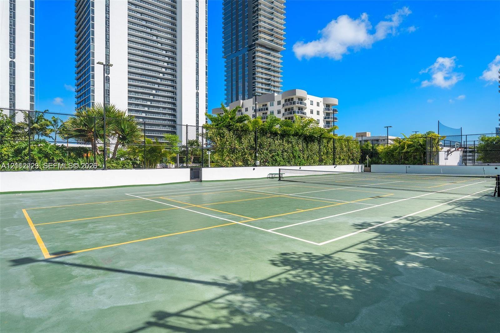 Tennis court featuring a specialized green surface, perimeter fencing, and surrounding mature landscaping
