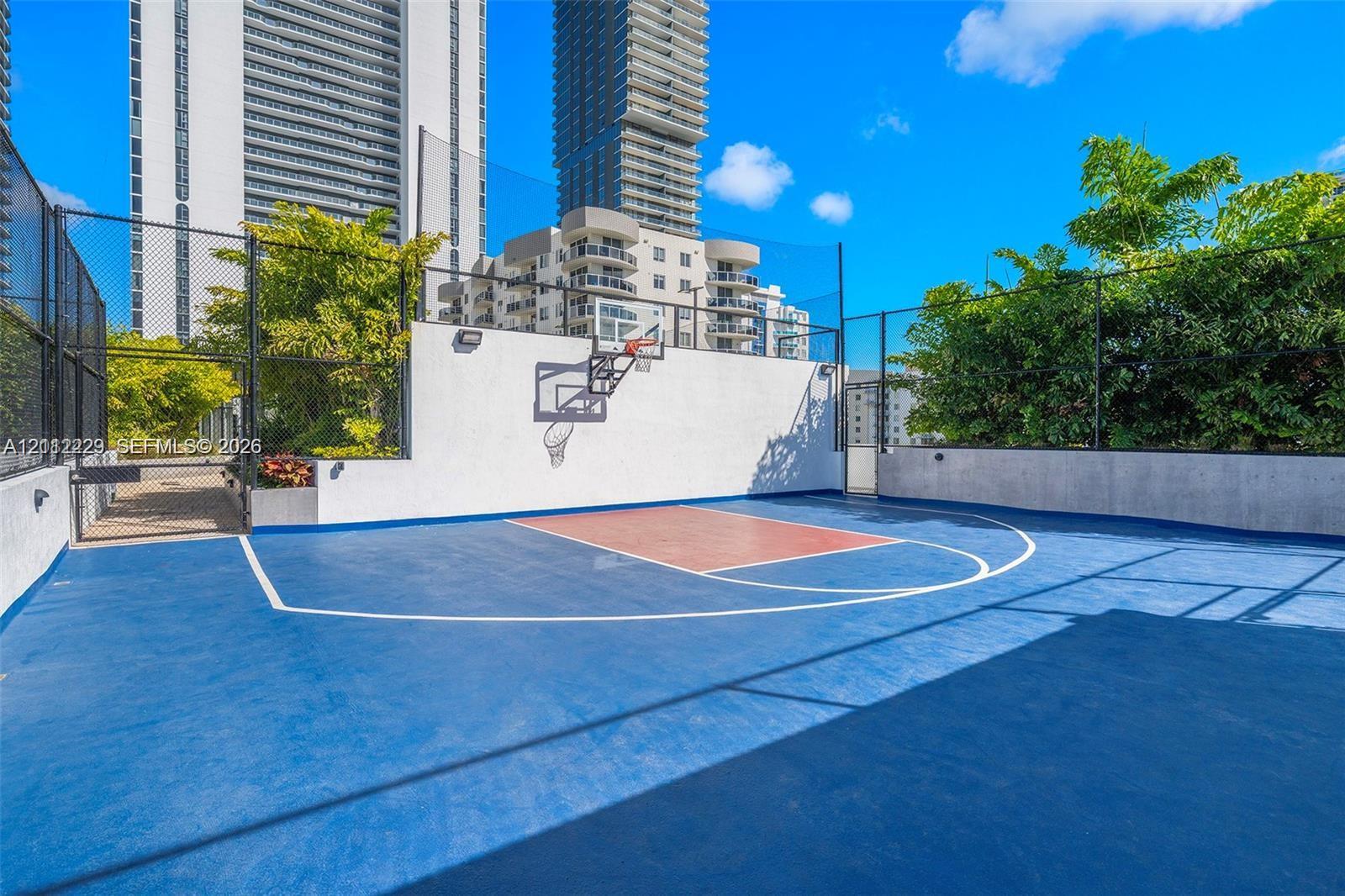 Outdoor basketball court featuring a multi-tone surface, wall-mounted hoop, and high perimeter mesh fencing