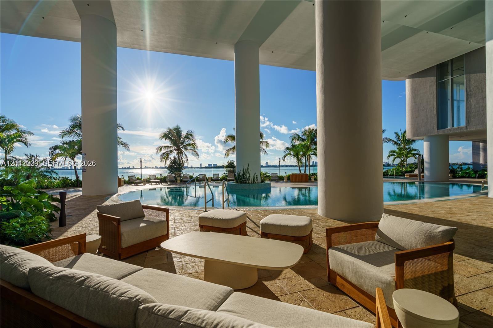 Covered outdoor terrace featuring stone-tiled flooring, prominent architectural columns, and a custom-shaped swimming pool overlooking the water