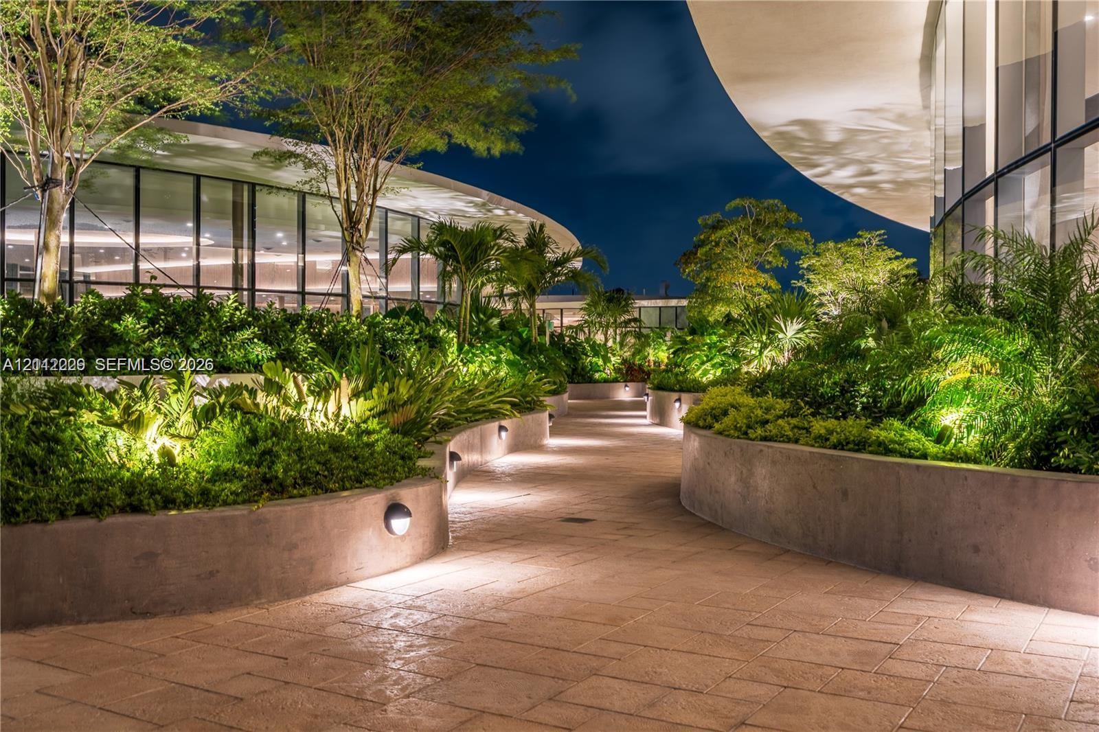 Curved exterior walkway featuring stone-tiled paving, tiered concrete retaining walls, integrated low-level lighting, and floor-to-ceiling glass panel walls